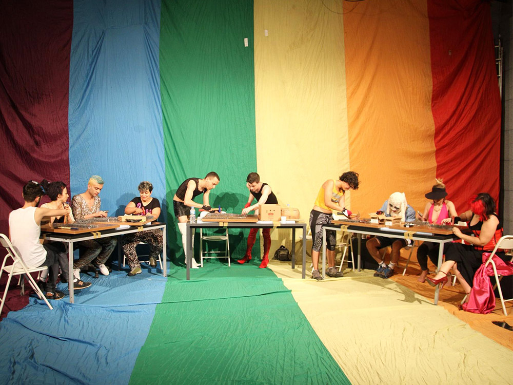 A group of people work at long tables in front of a rainbow flag that covers the back wall and the floor