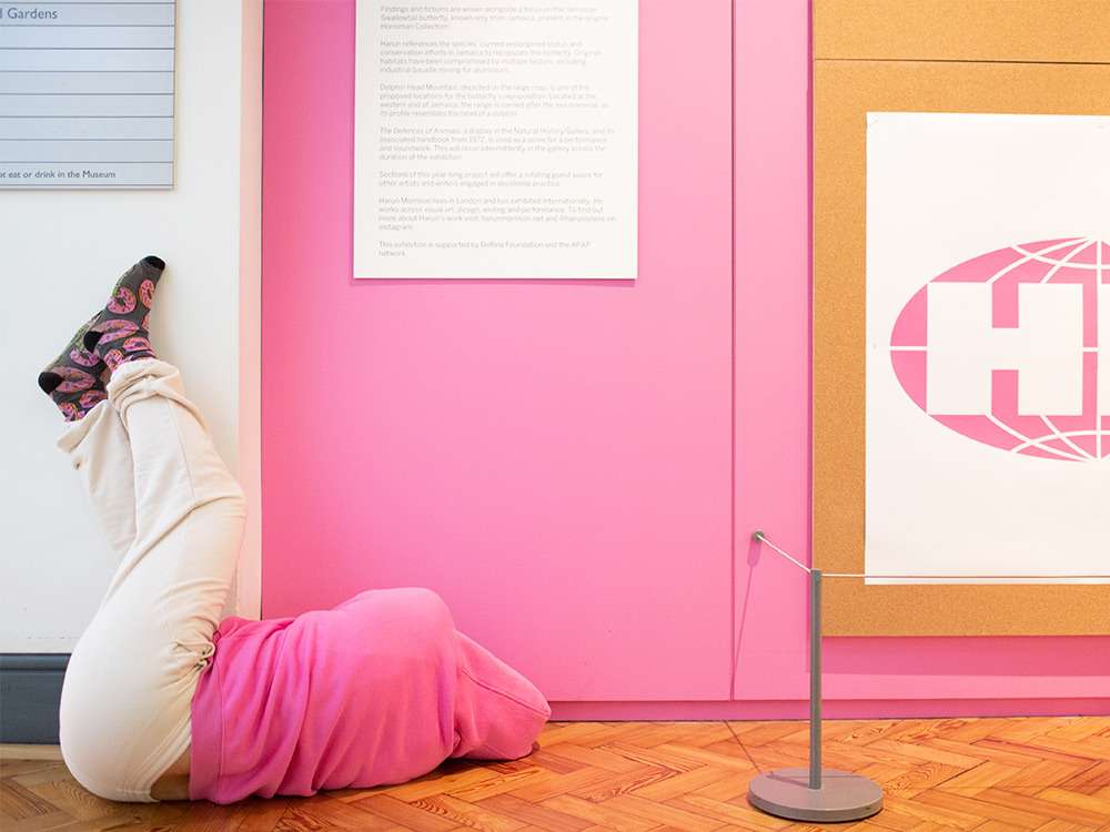 A figure in pink and white clothes blends into the wall, against a pink and white branded display of logos.