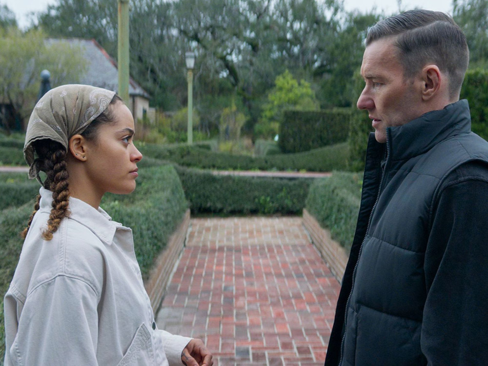 A young black woman and an older white man stare off at each other in front of meticulously kept hedges