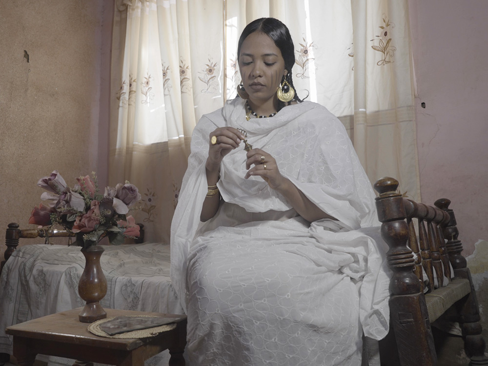 A person in a white flowing outfit holds a small artefact, sitting on their bed in their room