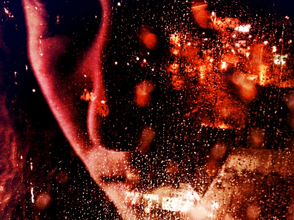 Close up photo of a person's face with their eyes closed, reflecting red light, traffic and light signs, behind glass covered by water droplets