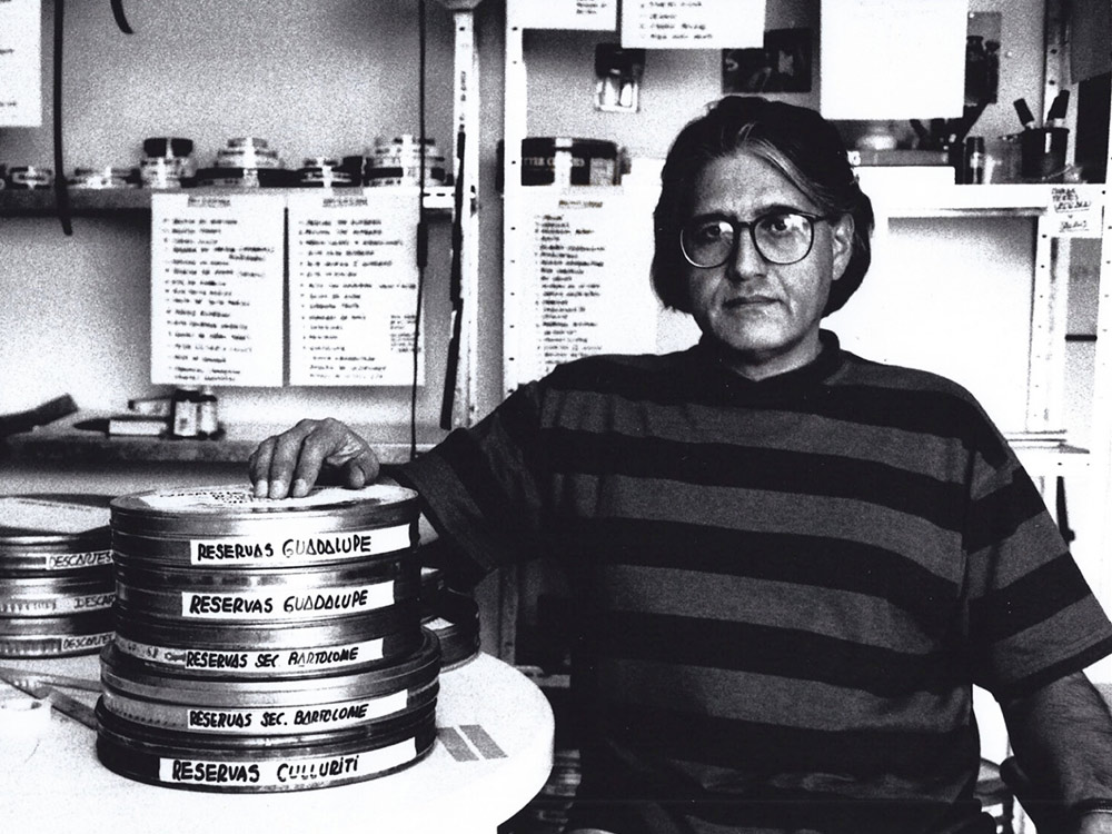 Filmmaker Patricio Guzmán sitting at a table, his hand on a stack of reels. The photo is in black and white. He's wearing round glasses and a striped tshirt