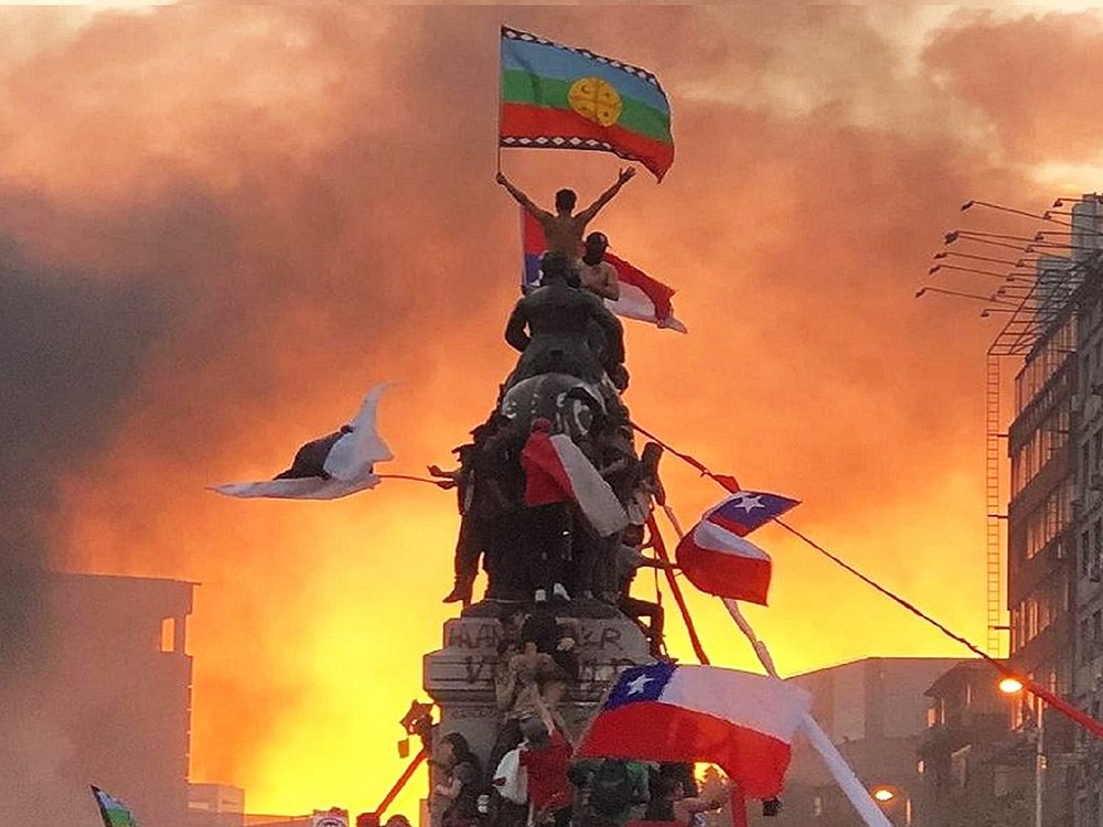 A statue is scaled by Chilean protesters against a smokey, fiery orange sunset