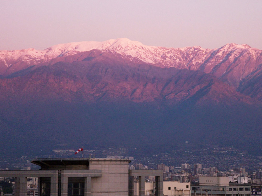 A huge snowcapped mountain range lies behind the tops of concrete buildings of a city