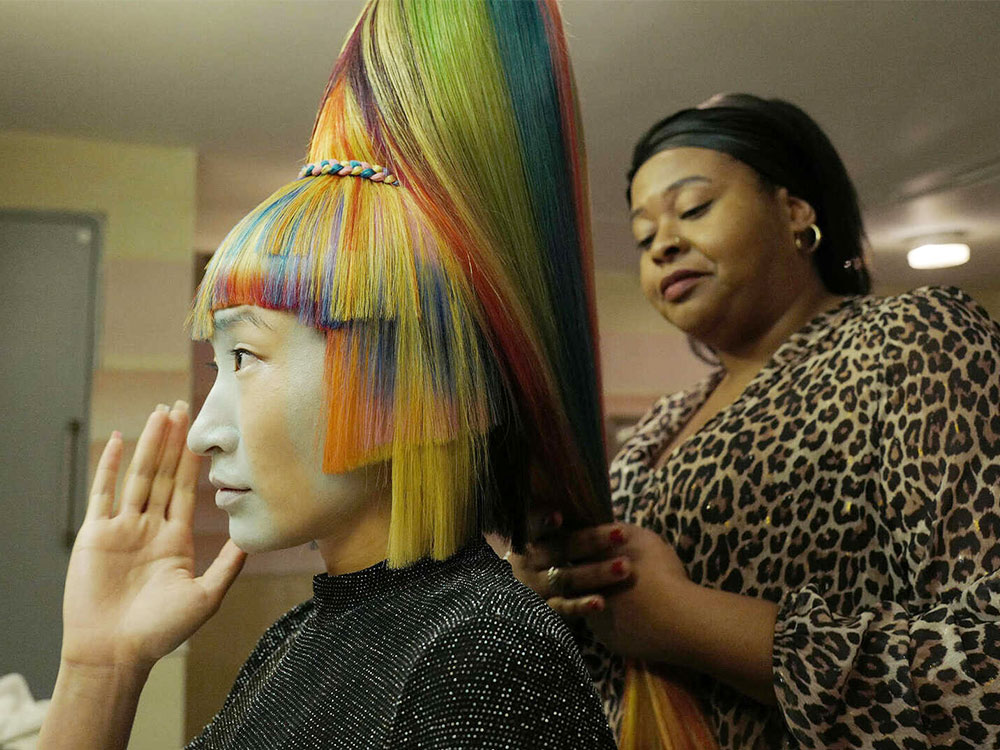 A black woman does the hair for an Asian person with rainbow-streaked hair and a white face