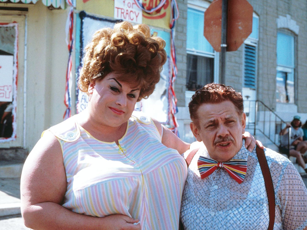 A woman with strong makeup and curls and a small man in overalls and rainbow bow tie stand side by side, on a suburban street corner