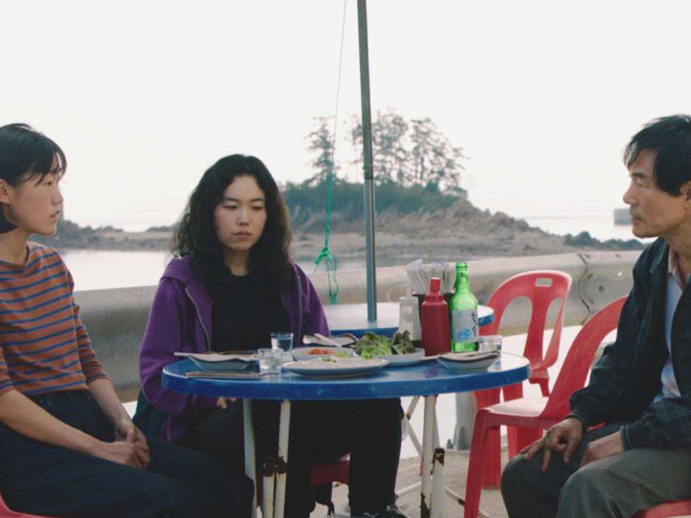 A Korean woman and her friend sit with her birth father on plastic chairs by the water, with soju and food