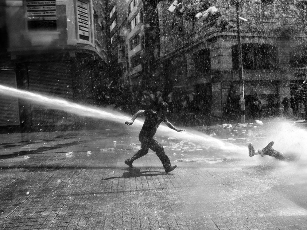 A black and white photo of a man running to aid a protester knocked down by a water cannon, on an empty street