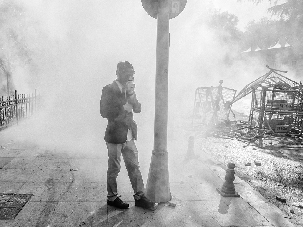 A man in a suit and gasmask stands by a lamp pole on a ruined street, covered in a cloud of dust