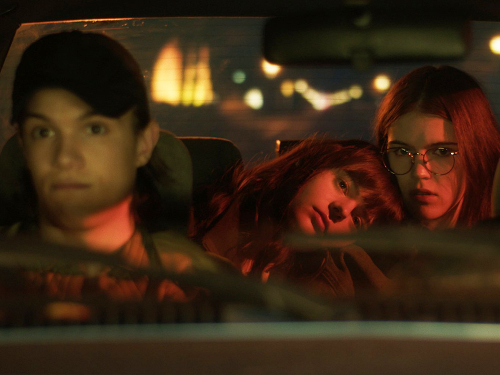 A young person in a cap drives a car. In the back, two young people, unsure of their journey