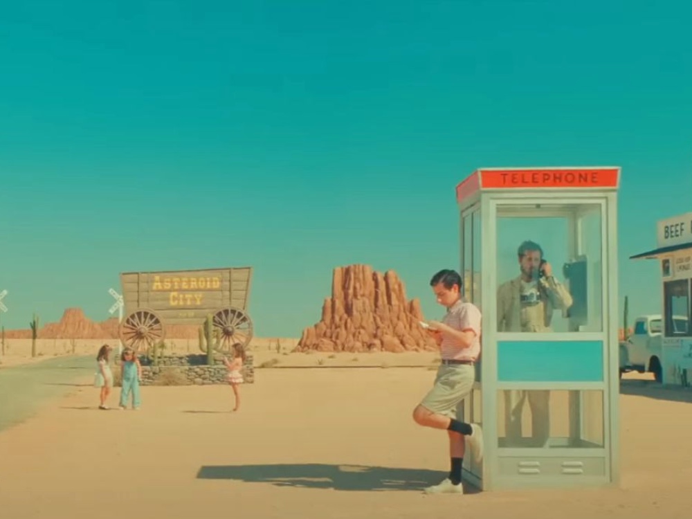 Painting-like desert landscape. Someone stands in a telephone booth and another leans against it. Children are standing in the background