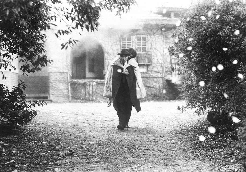 Black and white of someone being carried on someone's back through a garden, framed by bushes. They walk towards the camera, a building in the background.