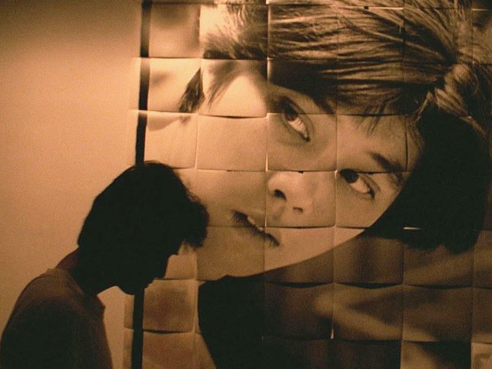 A short haired person is sat in shadow in front of a large portrait of someone. The portrait is made up of several equal sized squares. The whole image is sepia tone.