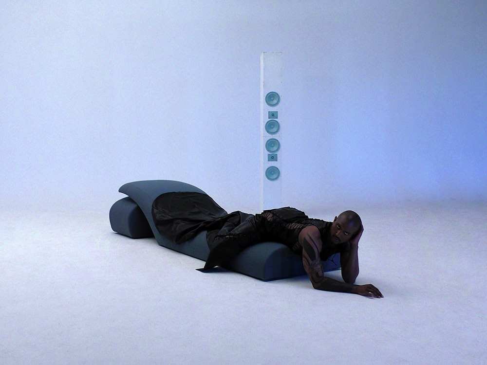LSDXOXO, a black man, wearing a long black dress, lying on a couch, with a single tall speaker stack floating behind him