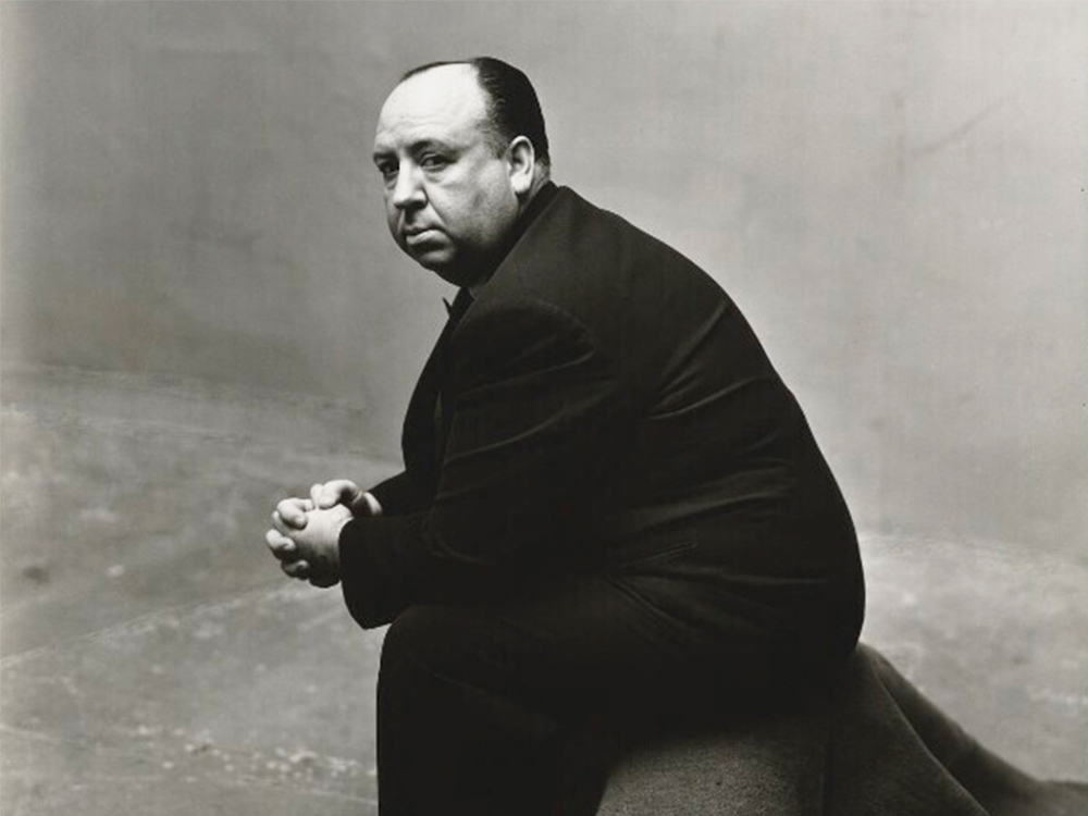 A portrait of Alfred Hitchcock, dressed in a suit, sitting side on, staring down the lens