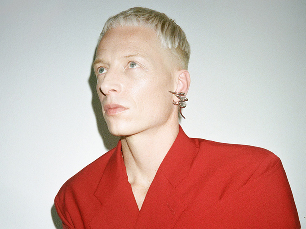 A headshot of Bendik Giske, a white person with short blonde hair, sharp earrings, and red clothing
