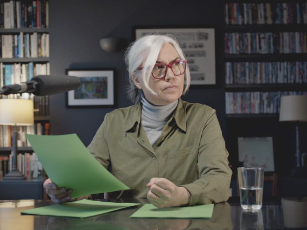 A person with grey hair, red framed glasses and a khaki shirt handles green papers. They're talking on mic, in a living room full of DVDs and books