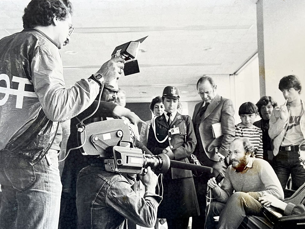 A man with a beard is being interviewed by a camera crew. A crowd looks on