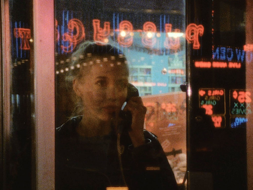 A person looks out of a window whilst on the phone, the reflections of neon lights are across their face