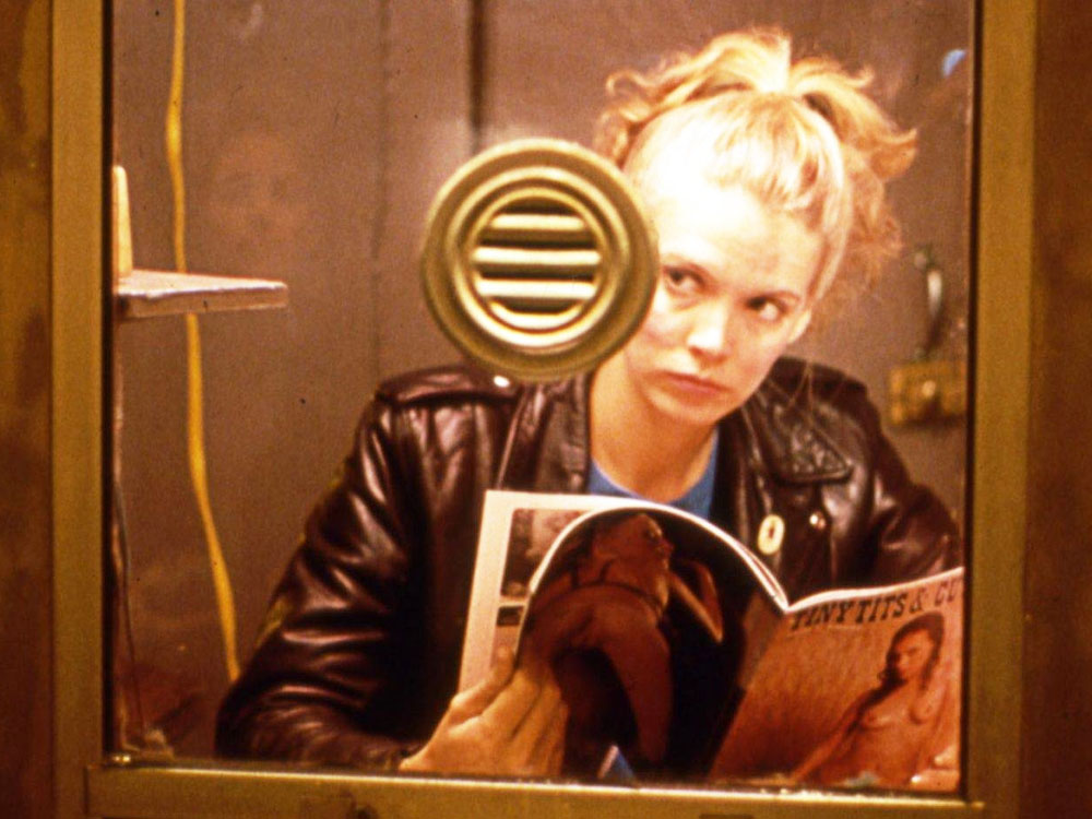 A blonde person in a leather jacket reads a porn mag behind a reinforced glass window. They're looking at someone, unseen