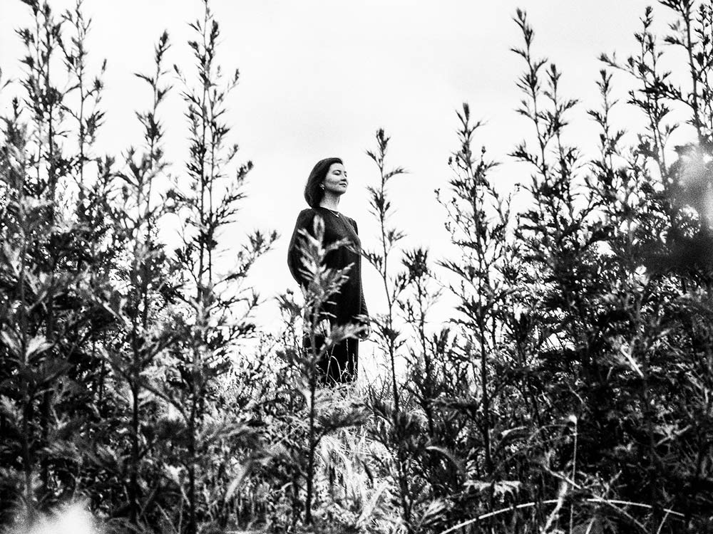 A black and white photo of Galya Bisengalieva standing amongst a field of wheat. Her eyes are closed, feeling breeze on her