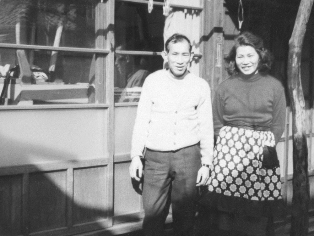 An elderly Japanese couple stand in front of a wooden house, smiling