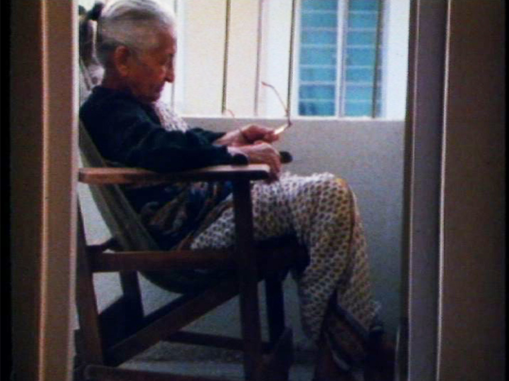 An elderly person sits in a balcony on their chair, glasses in hand
