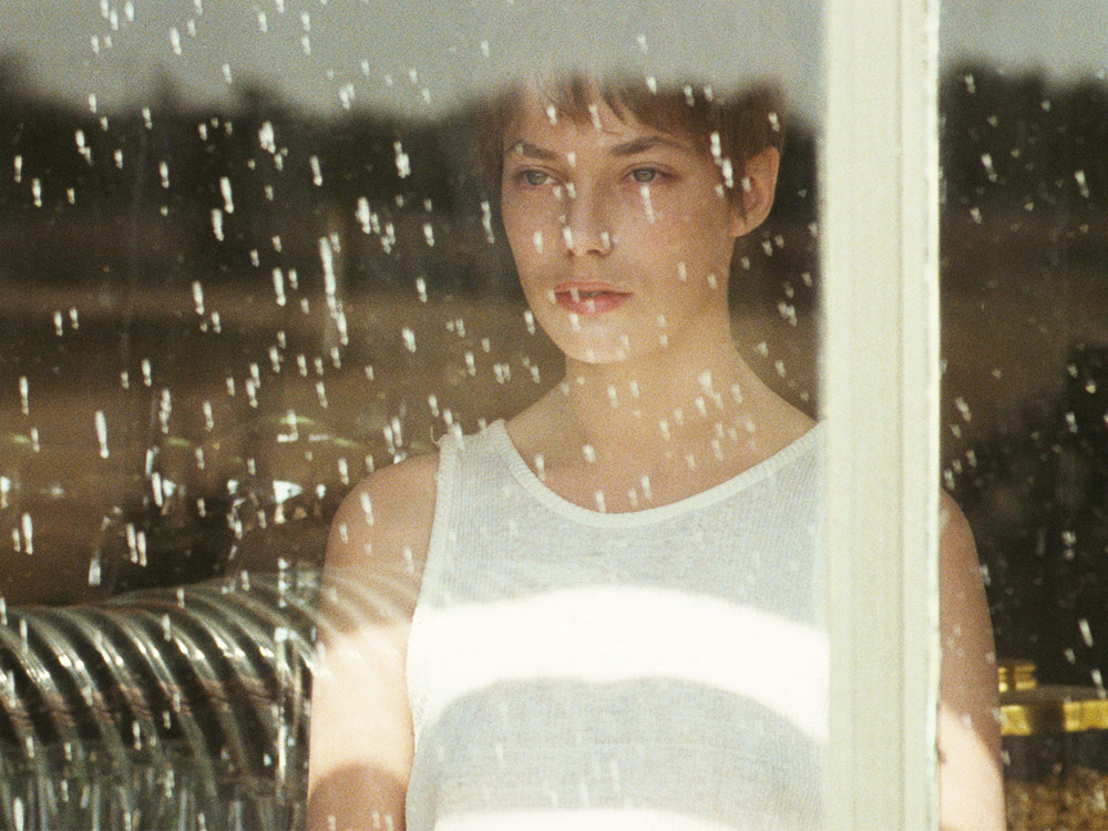 Jane Birkin, with short hair and a singlet, stares out a window at the rain