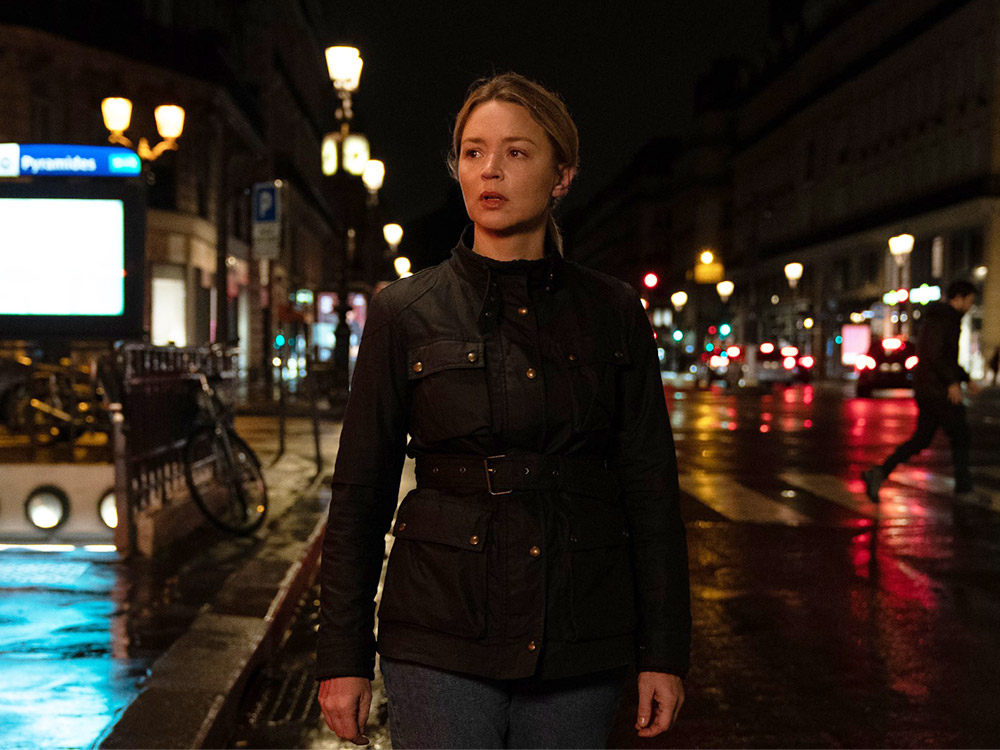 A blonde woman in a black overcoat walks the Paris streets at night