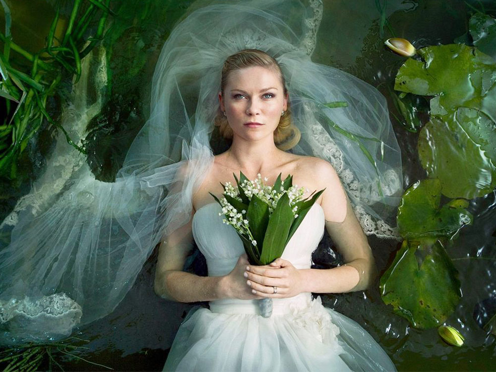A woman in a wedding dress holds a bouquet, lying in a shallow pool of water surrounded by reeds and lily pads. Her face is blank, staring skywards