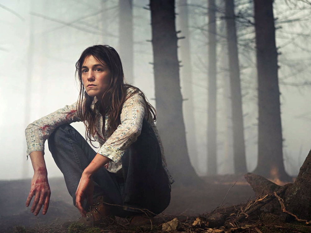 A woman with blood on her hands and clothes squats in a dark foggy forest, next to a broken tree