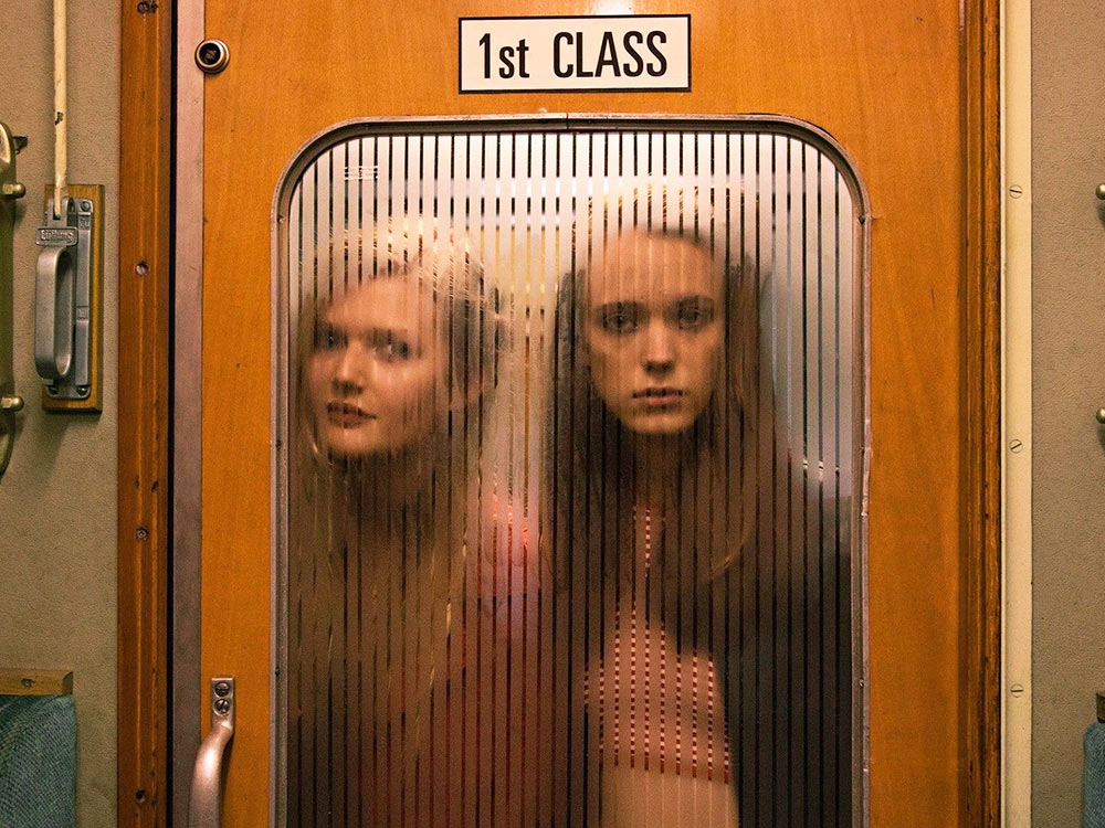 Two young women look through the window of a train cabin door marked '1st Class'