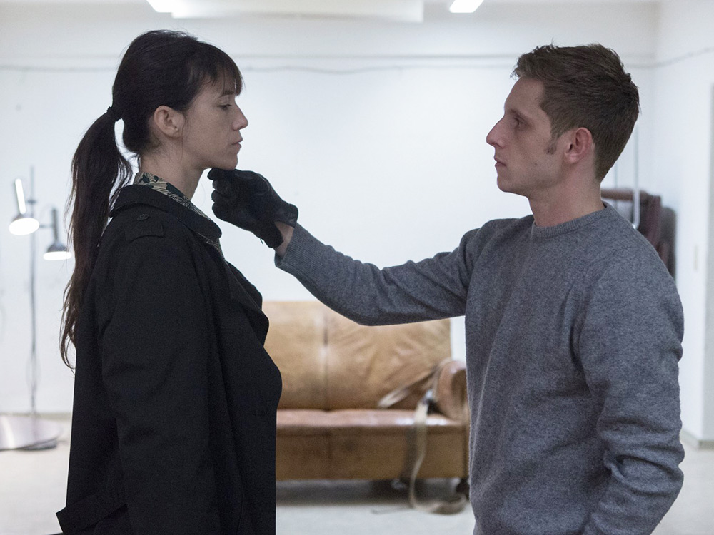 A man wearing gloves touches a women's chin, observing her face as she looks away