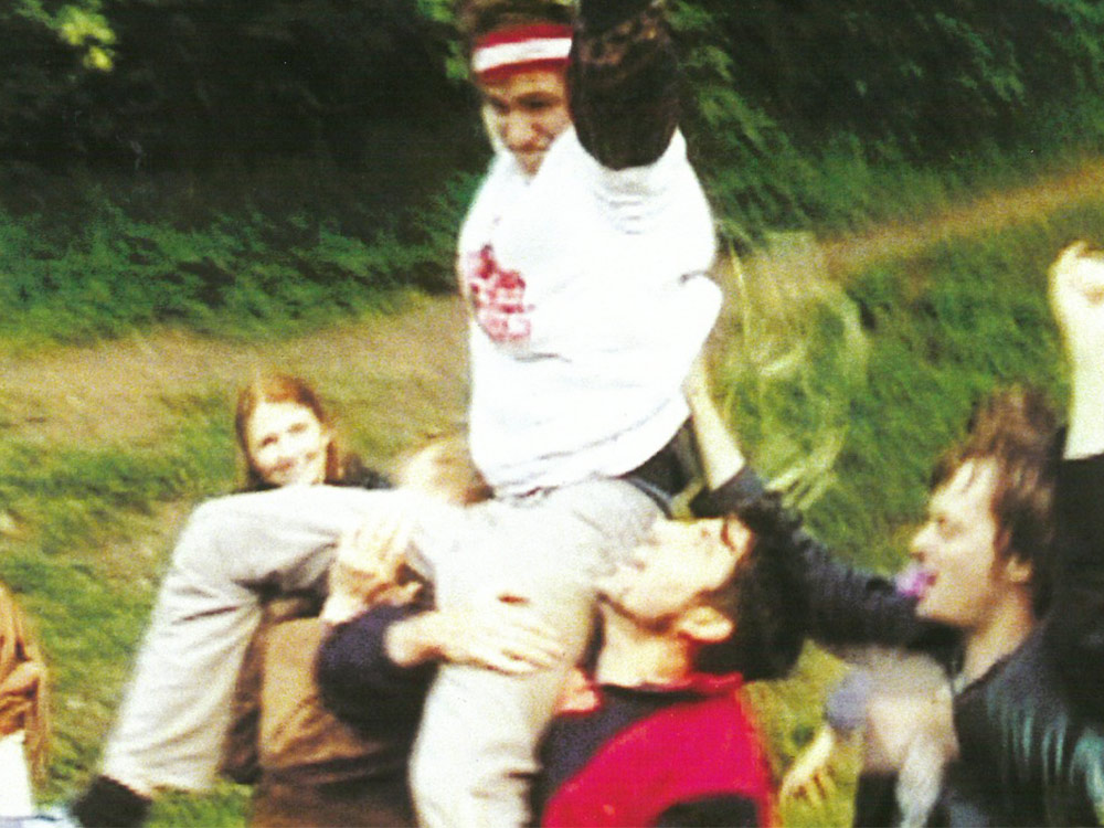 A group of young people walk through a field, cheering. One of them, wearing football gear, is sitting on top of their shoulders