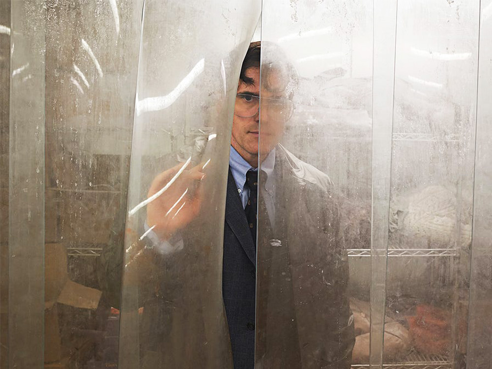 A man in a suit stares through grimy plastic curtain inside a freezer room