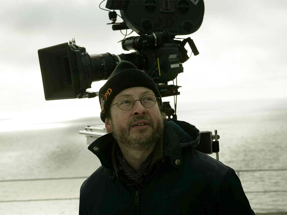 Director Lars von Trier in a beanie and winter coat. He's standing on a boat deck, overcast weather, with a film camera behind him