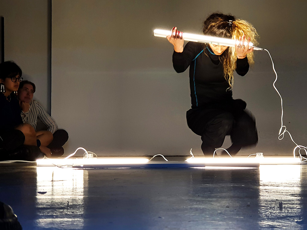 Artist Claudia Palazzo kneels on a hard floor lit by exposed fluoro lights. She is holding one, as if offering it