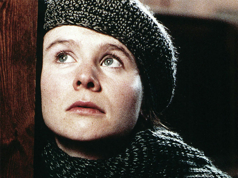 A person in a knitted woollen cap and top leans against a wooden surface, staring off