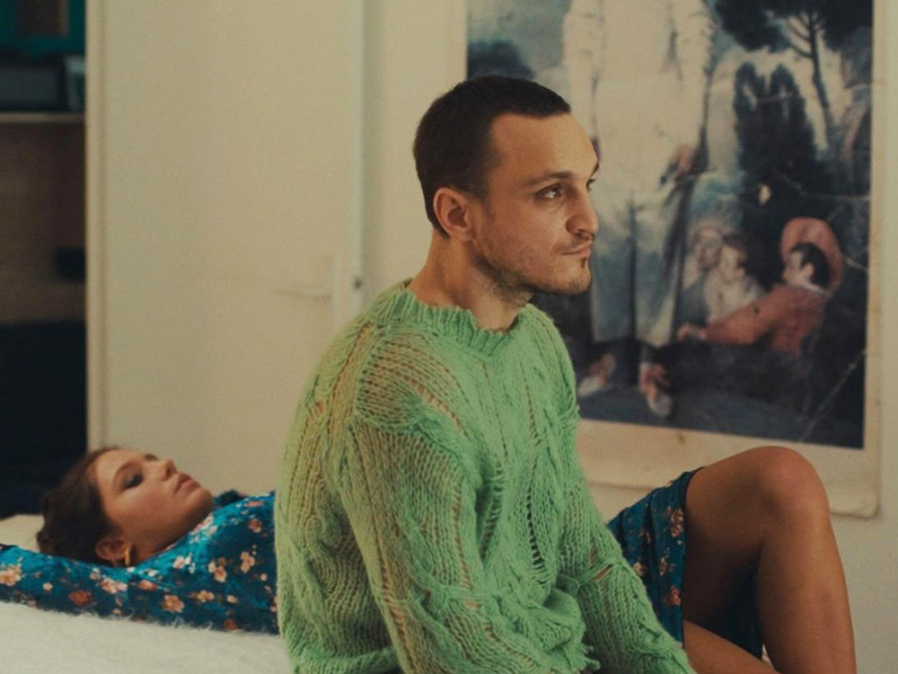A person with short hair and a green knitted jumper sits on a bed. Another person lies behind him, staring at the ceiling