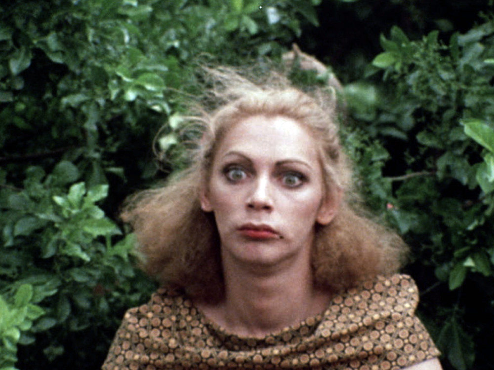 A person with messy blonde hair and wide eyes in front of bushes