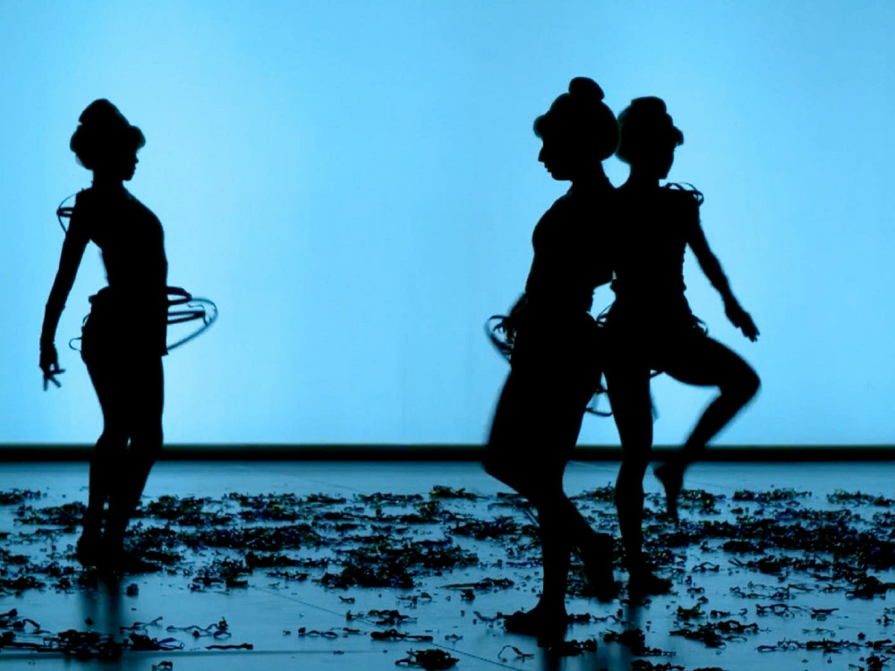 Dancers silhouetted against a bright blue screen. There is loose debree scattered across the floor, like ribbons