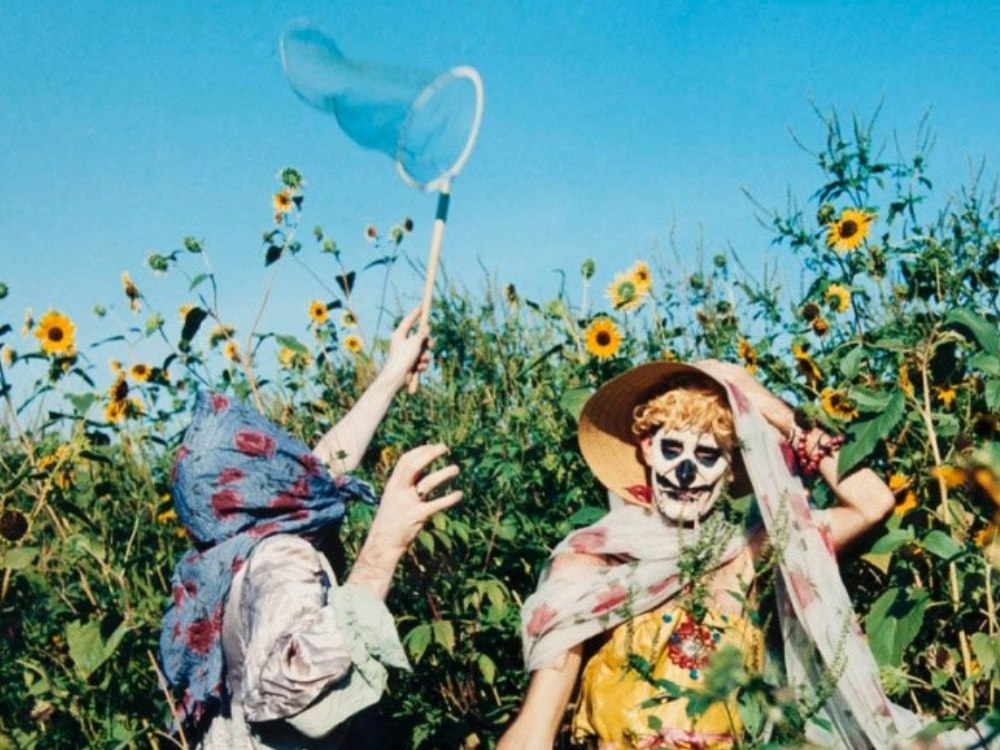 Two people in floral clothing stand in a field of sunflowers, blue sky, pulling a net through the air