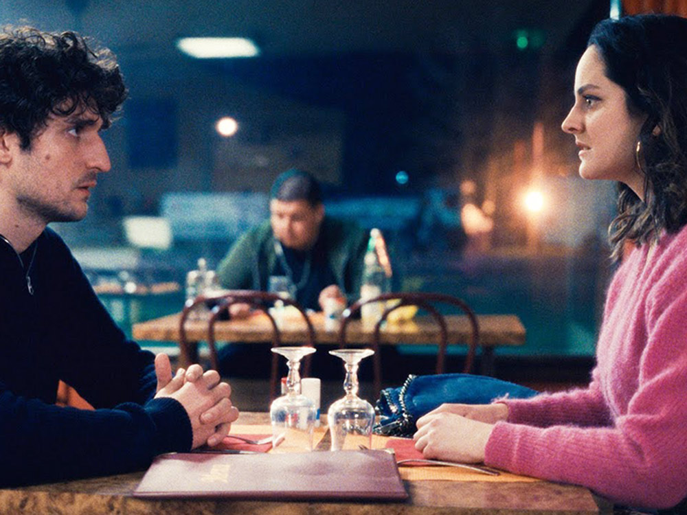 A man and woman sit opposite each other at a diner table, staring worriedly
