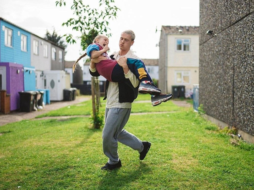 A young man carries a young girl playfully across the lawn of a housing block