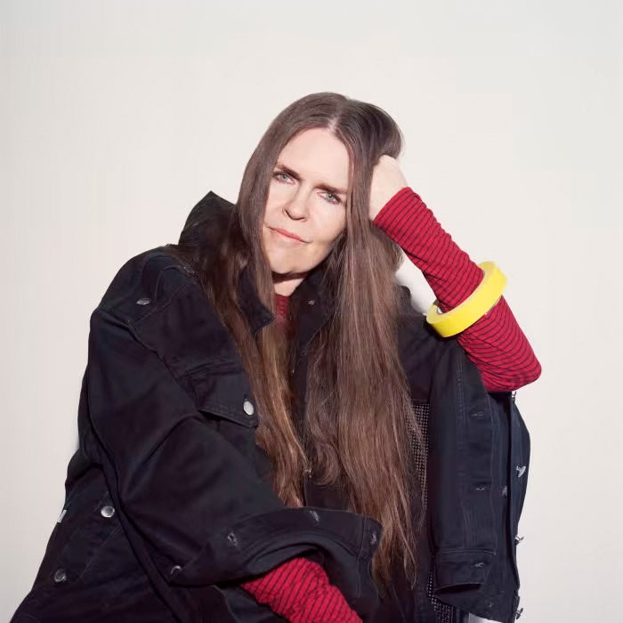A white person with long hair, wearing a striped red shirt, a black coat, and a yellow bangle around their arm