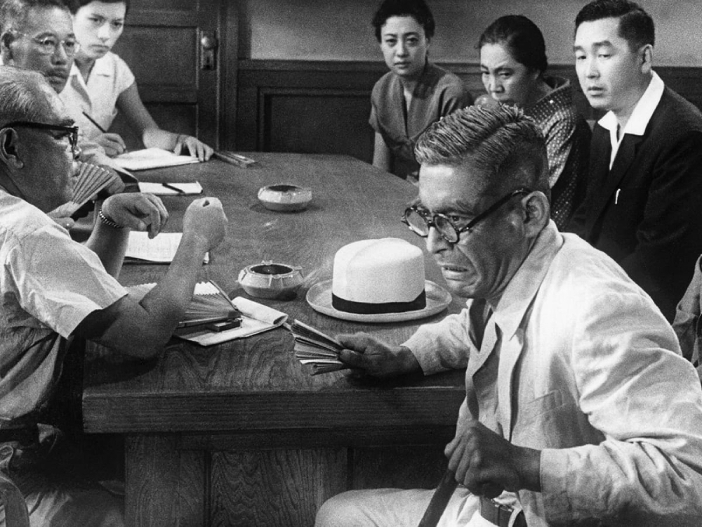 Seven Japanese people sit around a wooden table, the man in the foreground has a stressed expression. The image is in black and white