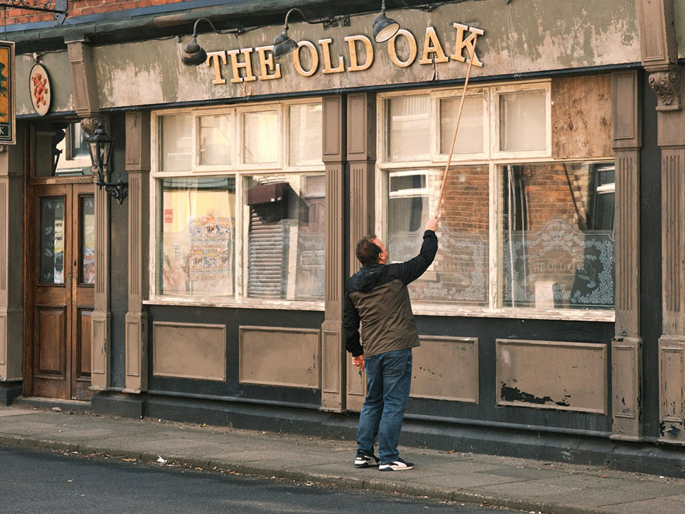 A white man stands in front of a run down pub, trying to adjust the wonky sign with a stick