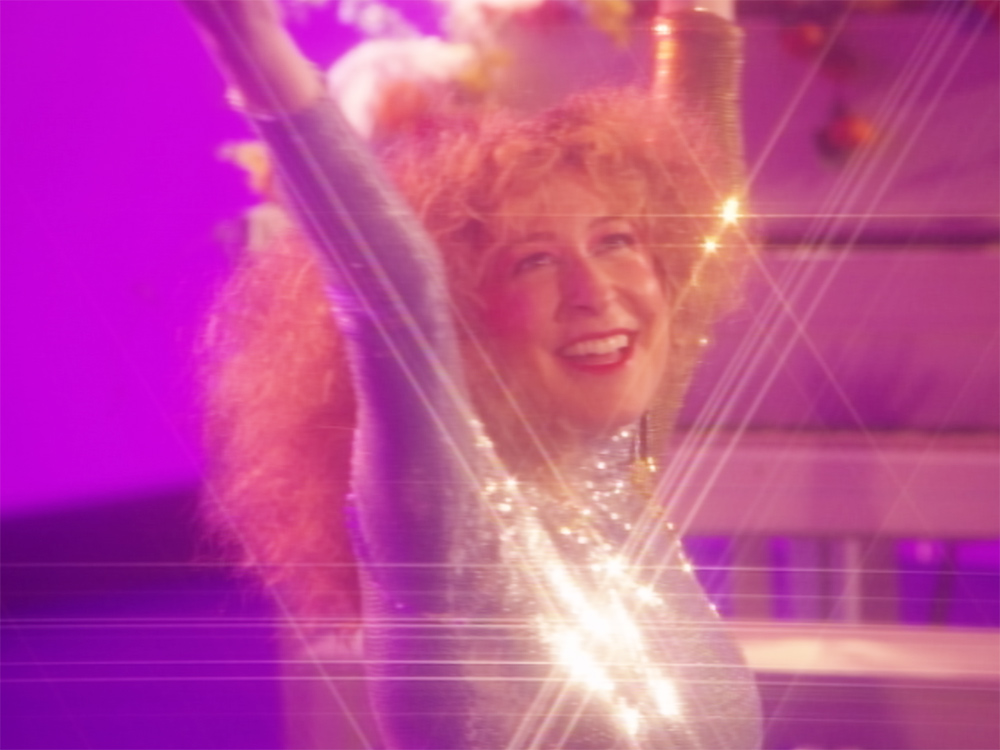 A blonde white woman in a sparkly top dances on a pink stage