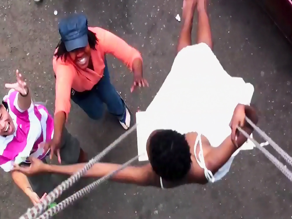 High angle of a black woman in a white dress swinging on a long rope swing, people below her grabbing at her and laughing