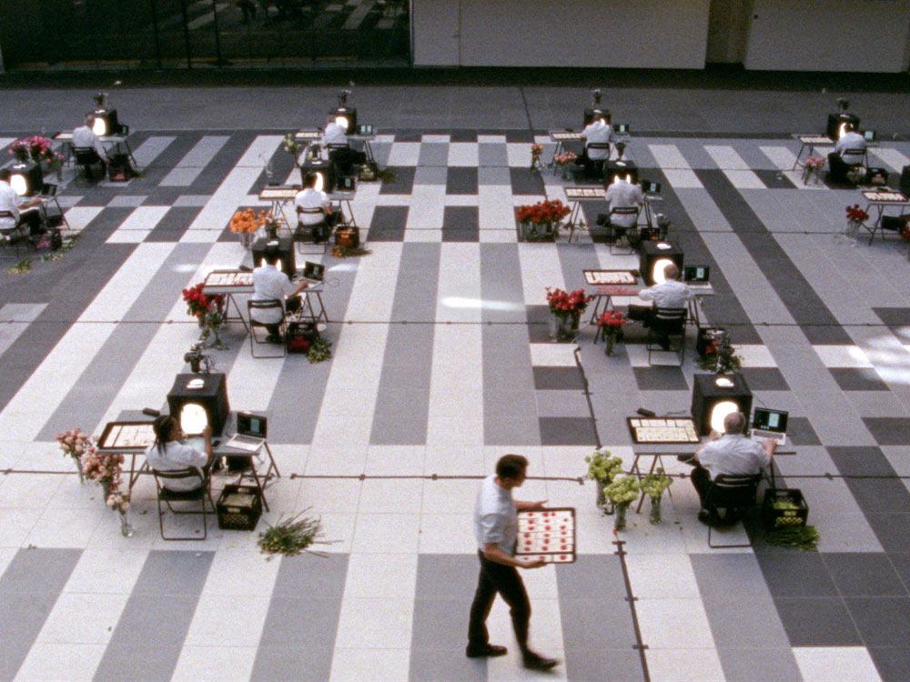 A room of men sit in a corporate-like building at their macbooks, but staring into black boxes full of light. A waiter walks around with small red food